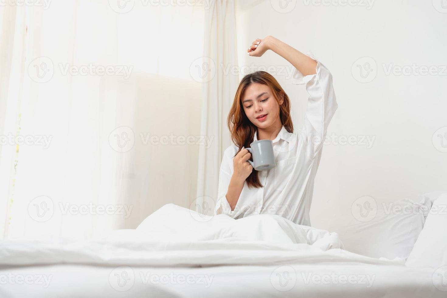 mujer en blanco bata de noche despertar arriba en fin de semana Mañana descansando y relajante jugando con ordenador portátil móvil teléfono comiendo un pan y Bebiendo té en vaso dentro blanco dormitorio. Mañana vacaciones concepto. foto