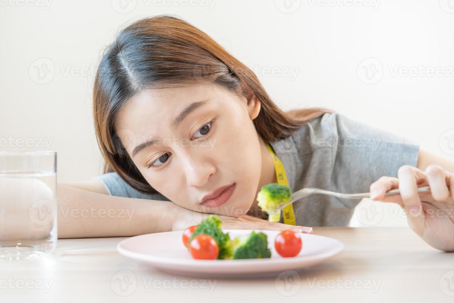 Anorexia, unhappy beautiful asian young woman, girl on dieting, holding fork at broccoli in salad plate, dislike or tired with eat fresh vegetables. Nutrition of good healthy, bored with food. photo