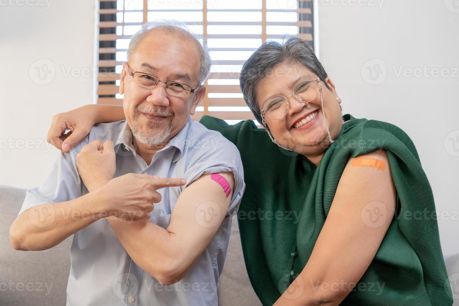 coronavirus vacunación, contento asiático anciano, Envejecido familia sonrisa fuerte juntos, demostración vendaje en brazo con proteger de covid-19 después inyección de vacuna, sentado en sofá en vivo habitación a hogar. foto