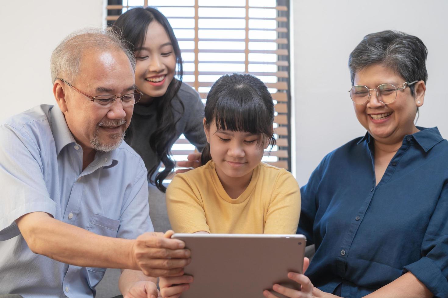 Happy family asian aged grandfather, grandmother have fun, smiling parent, girl using touchpad watching funny video call, play games and take photo selfie enjoy weekend together in living room at home