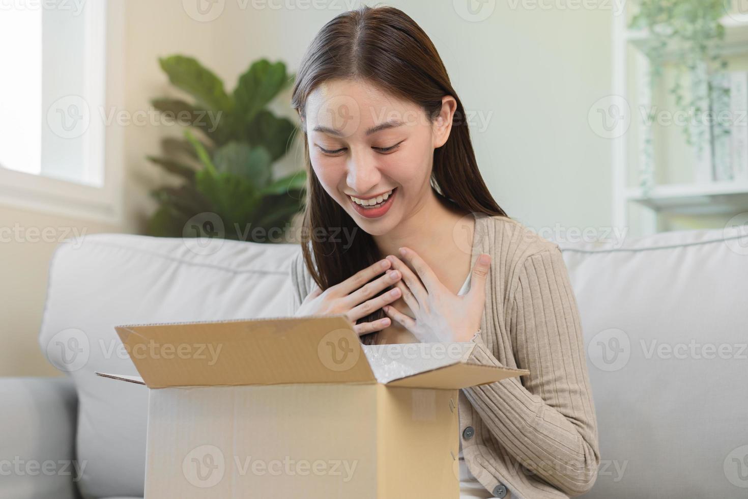 Happy excited, asian young woman, girl customer sitting on sofa at home, opening and unpacking cardboard box carton parcel after buying ordering present, shopping online, delivery service concept. photo