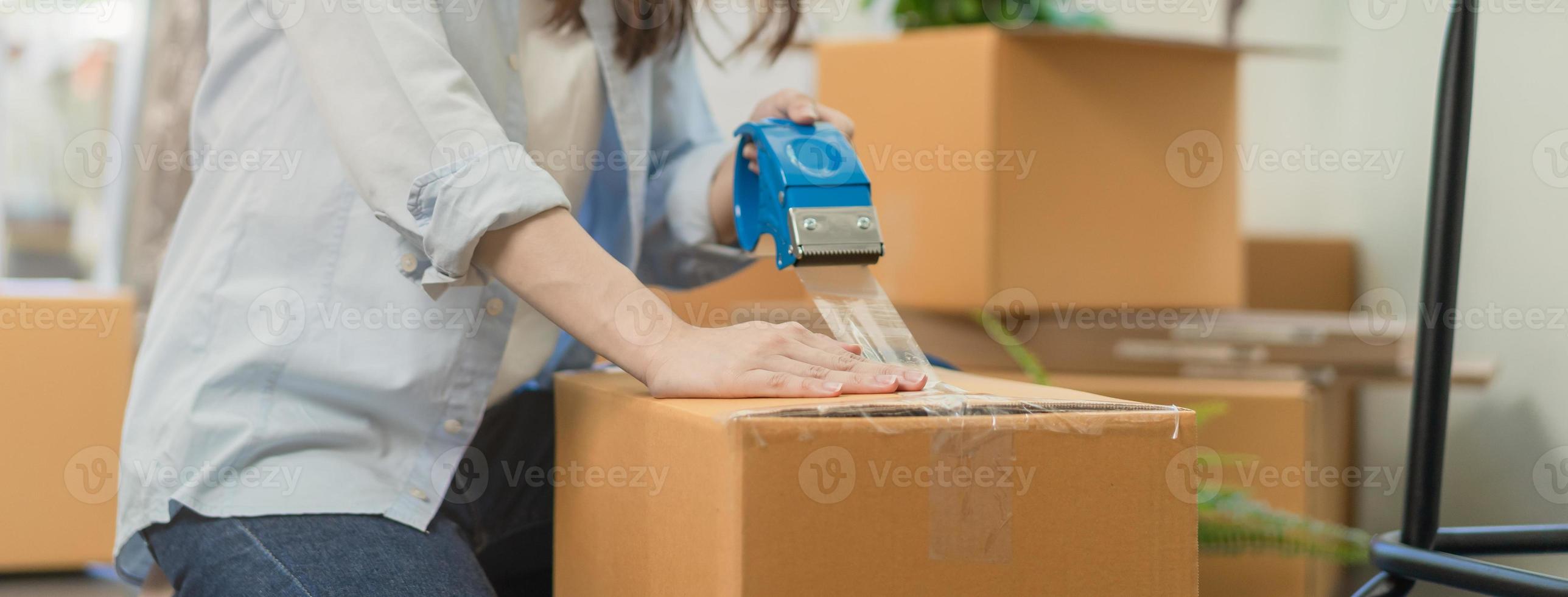 Focus hand of owner business asian woman, female holding packing box by tape machine, sealing cardboard indoors. Business owner in warehouse, Moving or preparing in new house, apartment or relocation. photo