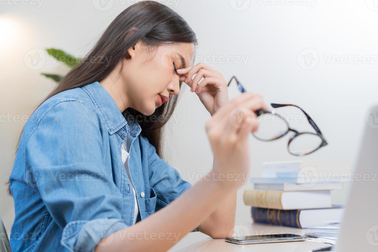 Exhausted, stressed asian young business woman working, took off glasses, massage nose from dry eyes, suffer on hard work while use laptop computer at office home. Overtime job, debt problem people. photo