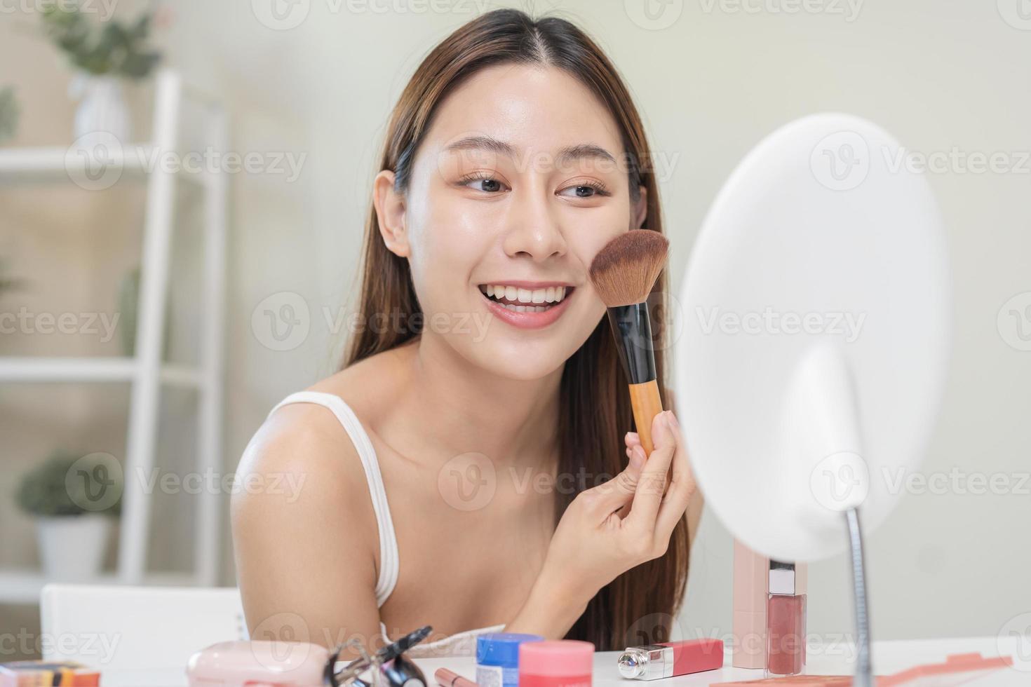 contento belleza blogger concepto, linda asiático joven mujer, niña sonrisa, hacer arriba cara por aplicando cepillo sonrojo polvo en su mejilla, mirando a el espejo. personas Mira con natural Moda estilo. foto
