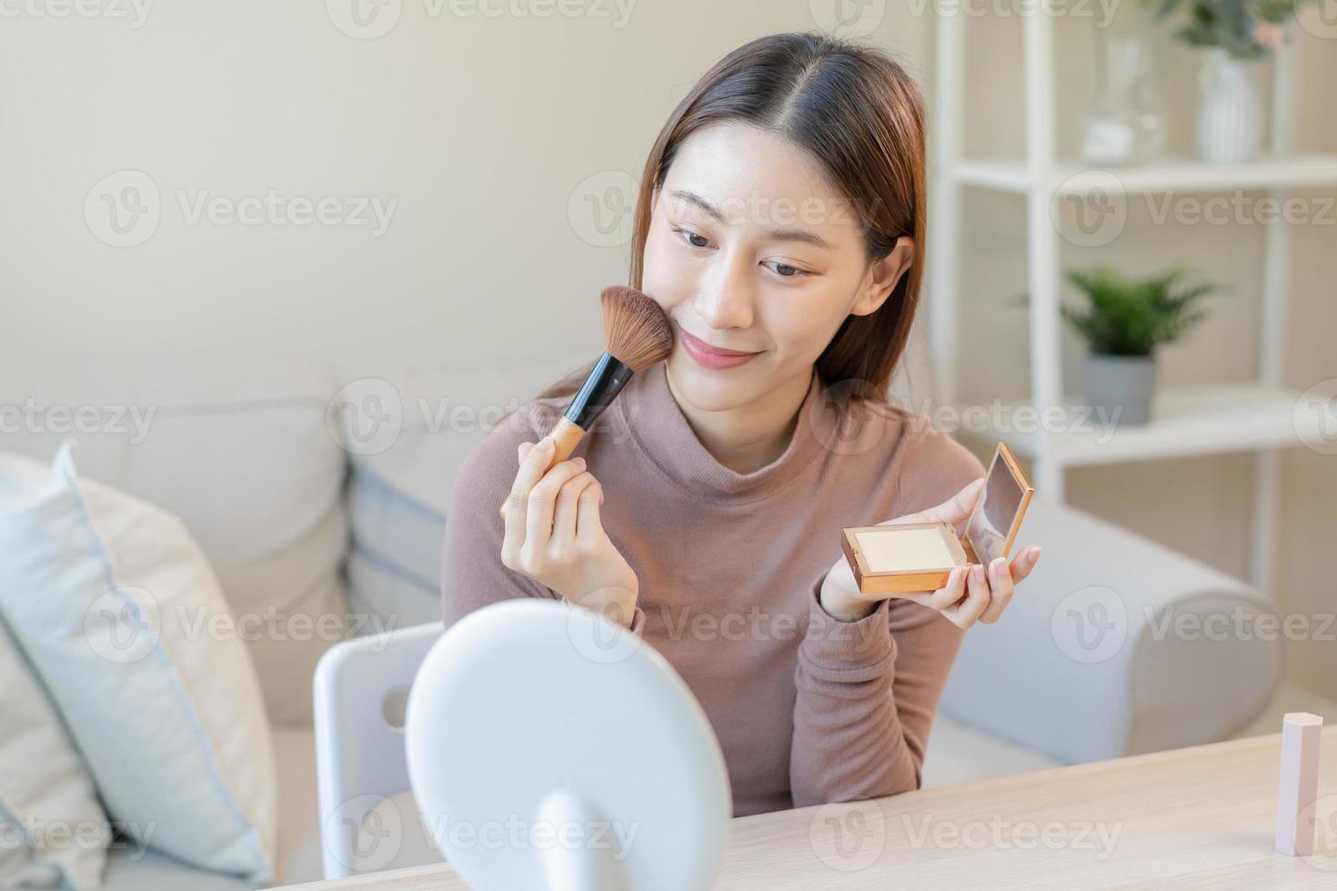contento belleza blogger concepto, bonito asiático joven mujer, niña hacer arriba cara por aplicando polvo Fundación por cepillo alrededor rostro, mirando a el espejo a hogar. hembra Mira con natural Moda estilo. foto