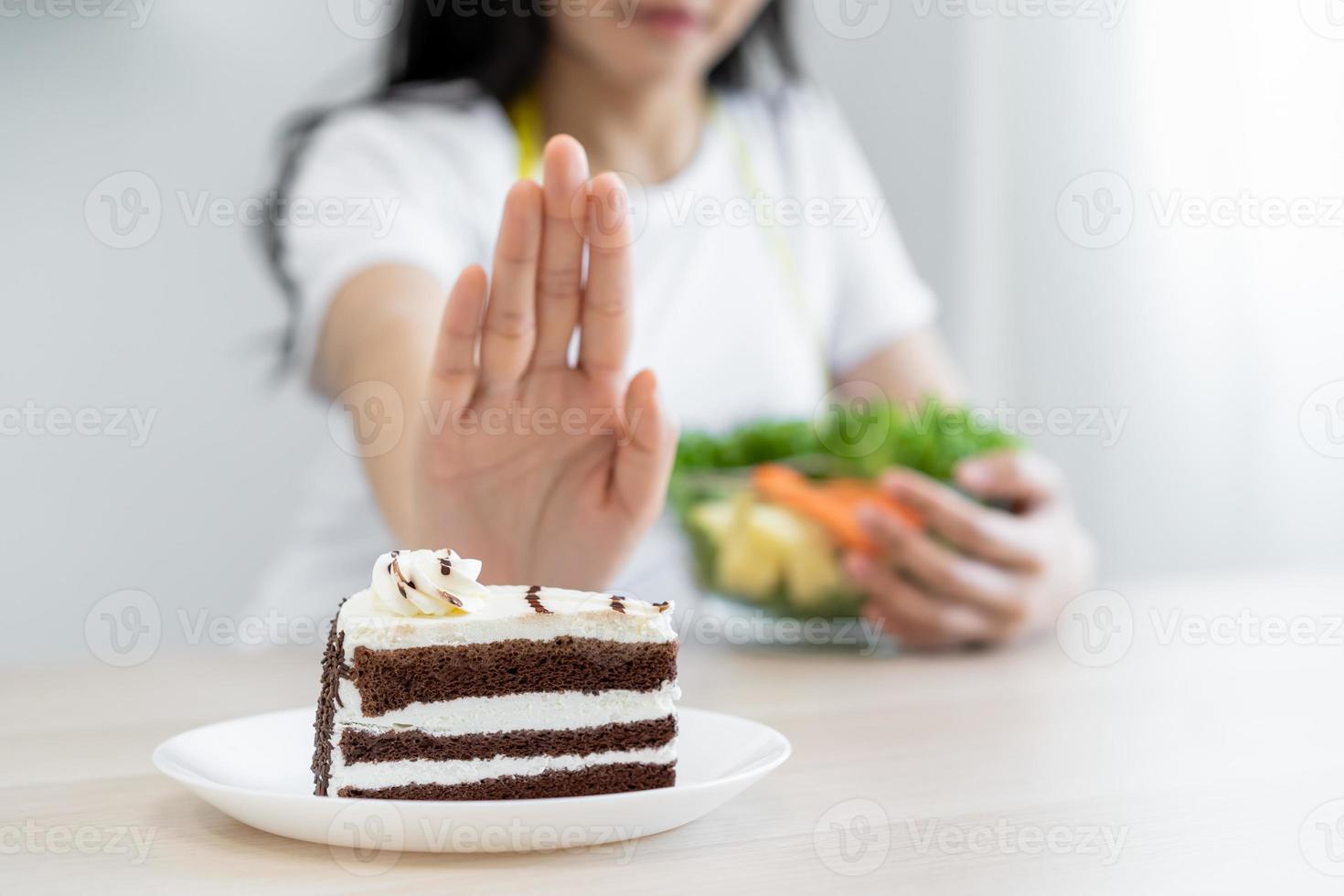 Diet, Dieting asian young woman using hand push out, deny chocolate cake or sweet taste, dessert food, choose eat green salad vegetables when hungry. Nutritionist of healthy, nutrition of weight loss. photo