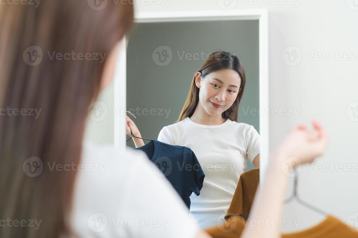 Choice of clothes,Nothing to wear. Attractive asian young woman, girl looking into mirror, try on appare, choosing dress, outfit on hanger in wardrobe at home. Deciding blouse what to put on which one photo