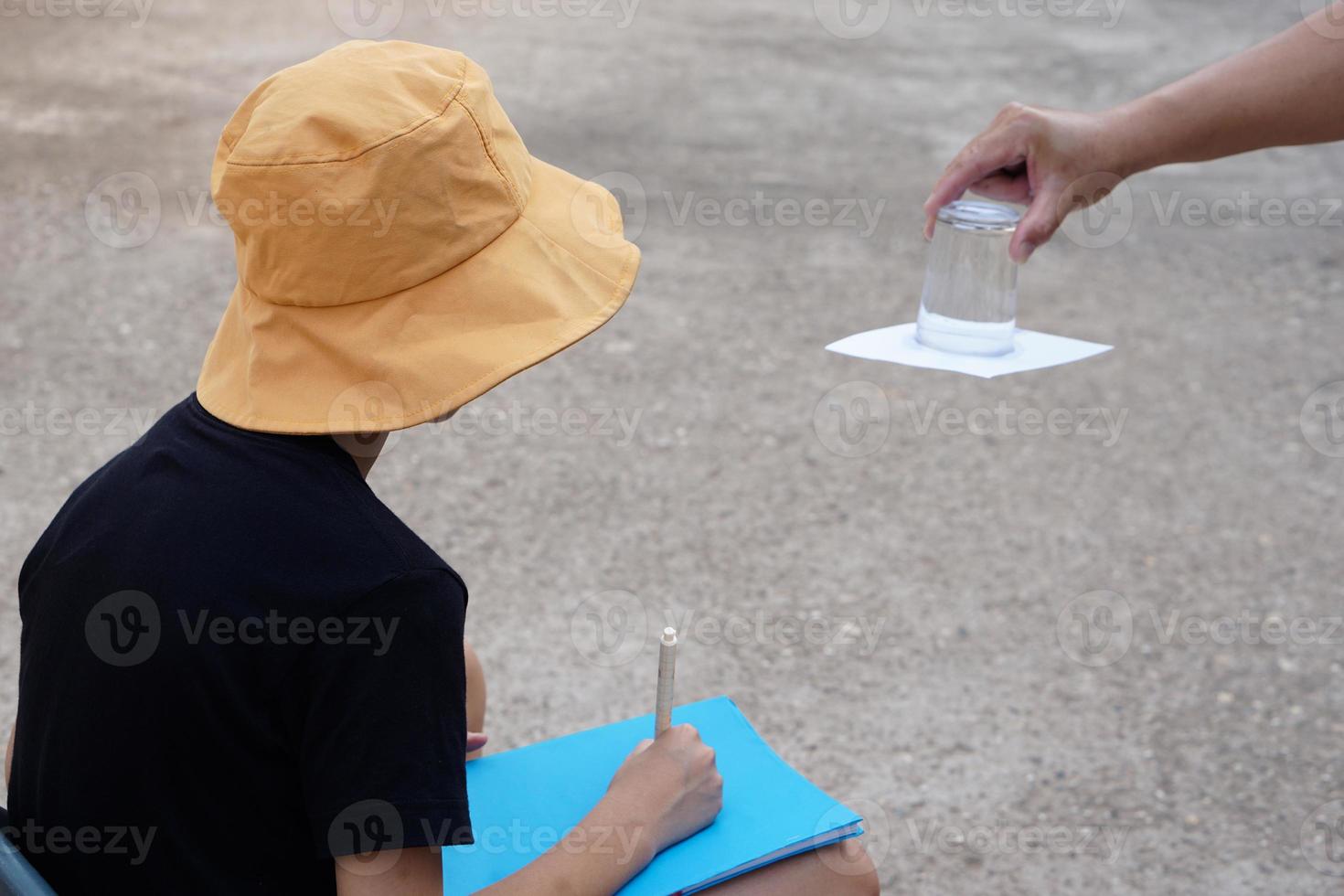 Student boy wears hat, hold book, learning science experiment about air pressure from glass of water which covered by paper. Concept, science subject activity, education. Learning by doing photo