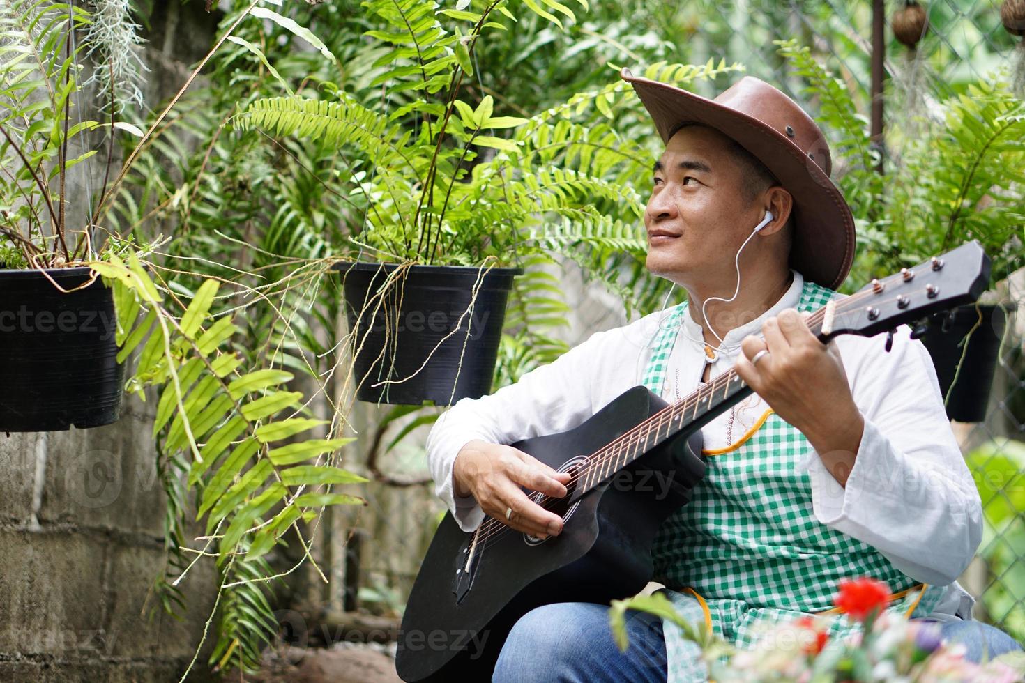 Asian man is playing the acoustic guitar in his home back yard garden. Concept, recreation activity, relax time with music and nature. Hobby, pastime. Love song and music. Easy living lifestyle. photo