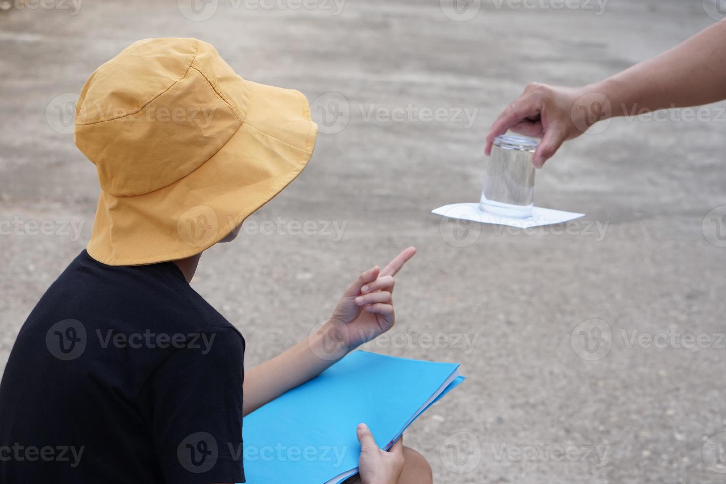 Student boy wears hat, hold book, learning science experiment about air pressure from glass of water which covered by paper. Concept, science subject activity, education. Learning by doing photo