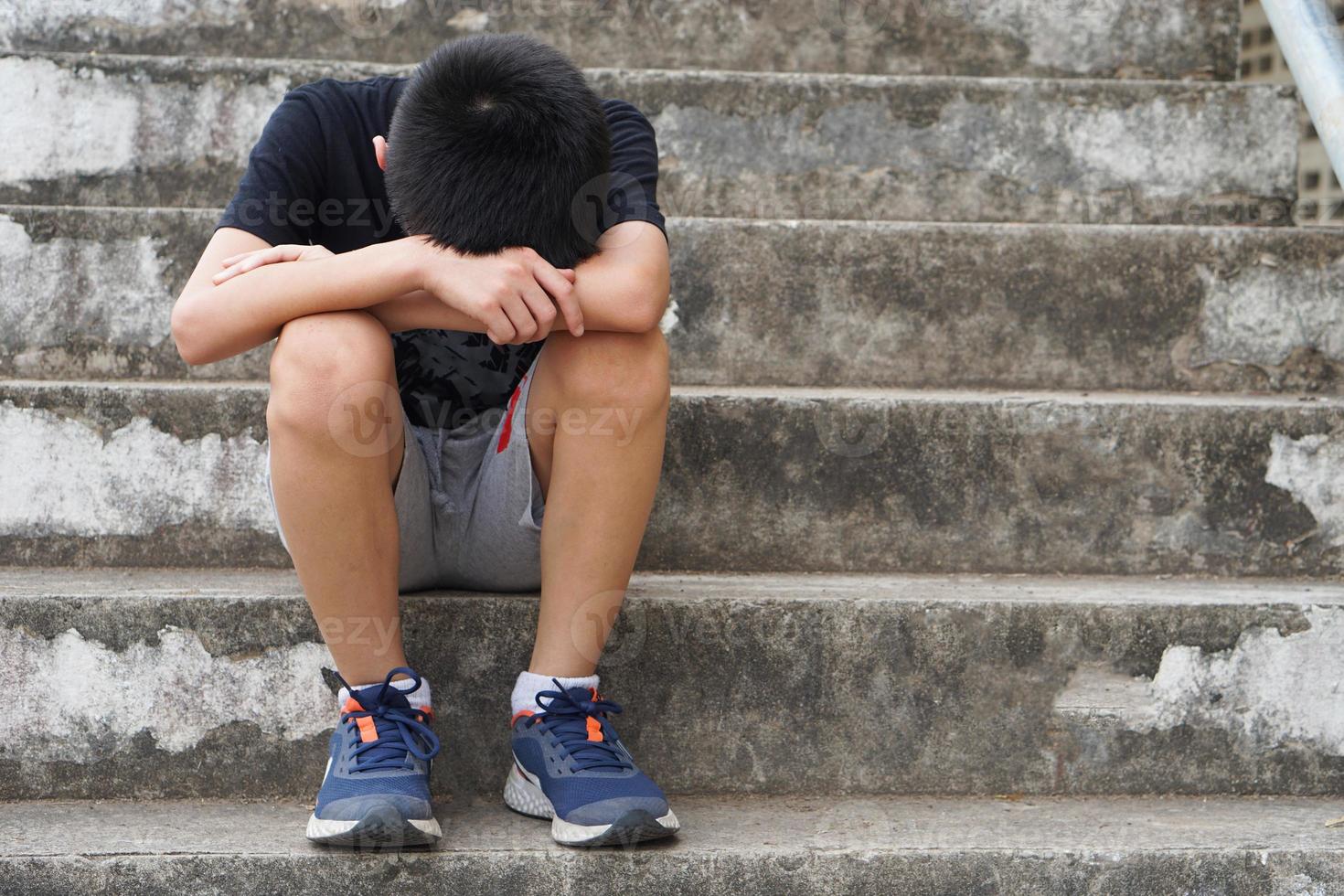 Depressed boy sits alone outdoor, sad and cry, face down between his knees. concept, emotion, feeling. Disappointed unhappy guilty . Boy is bullied and punished. Lonely. photo