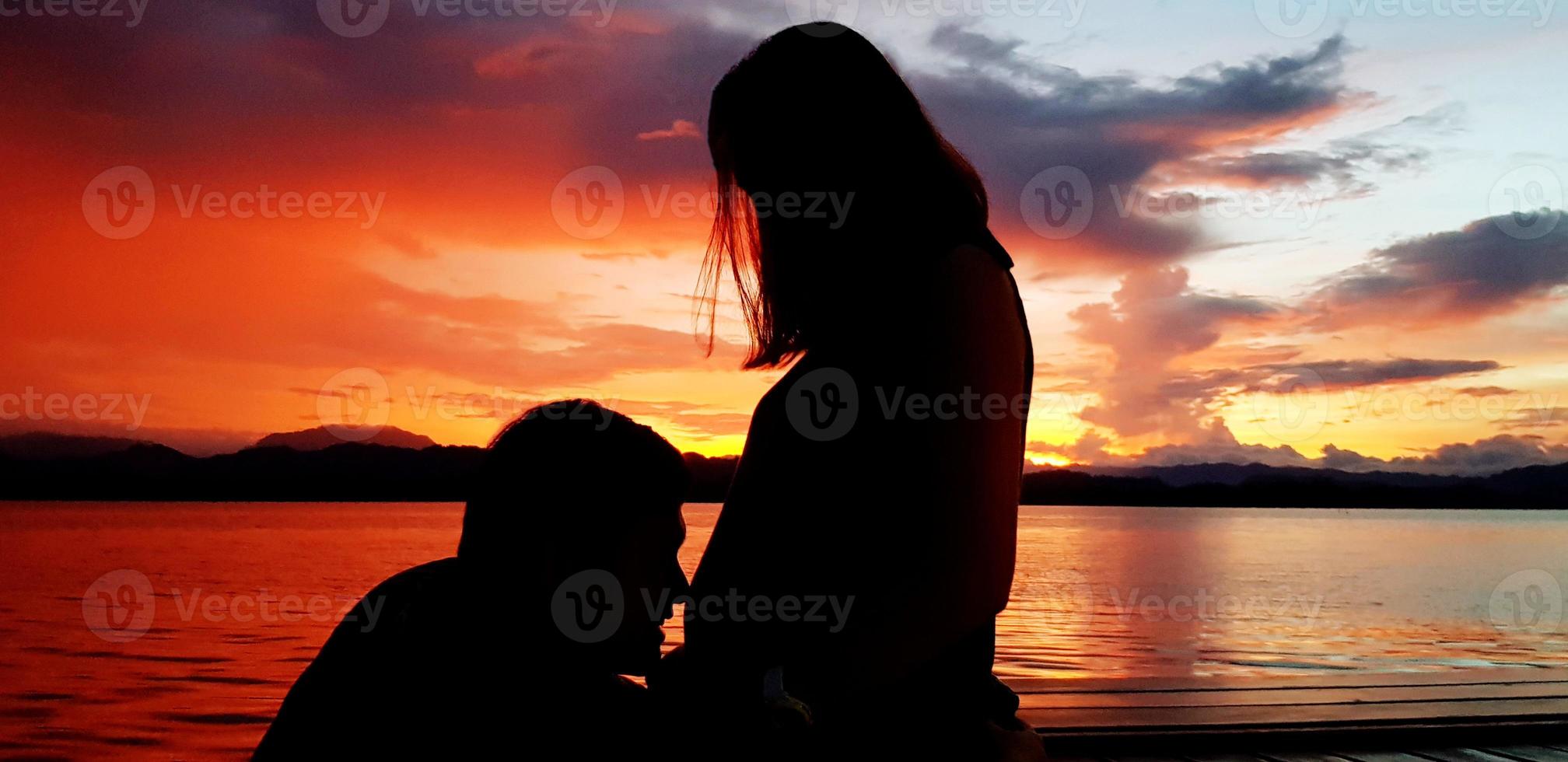 Silhouette of couple lovely at sunset time. Husband kiss on wife's belly there has baby inside with orange sky, cloud and lake background. Lover of family or parents and Pregnant concep photo