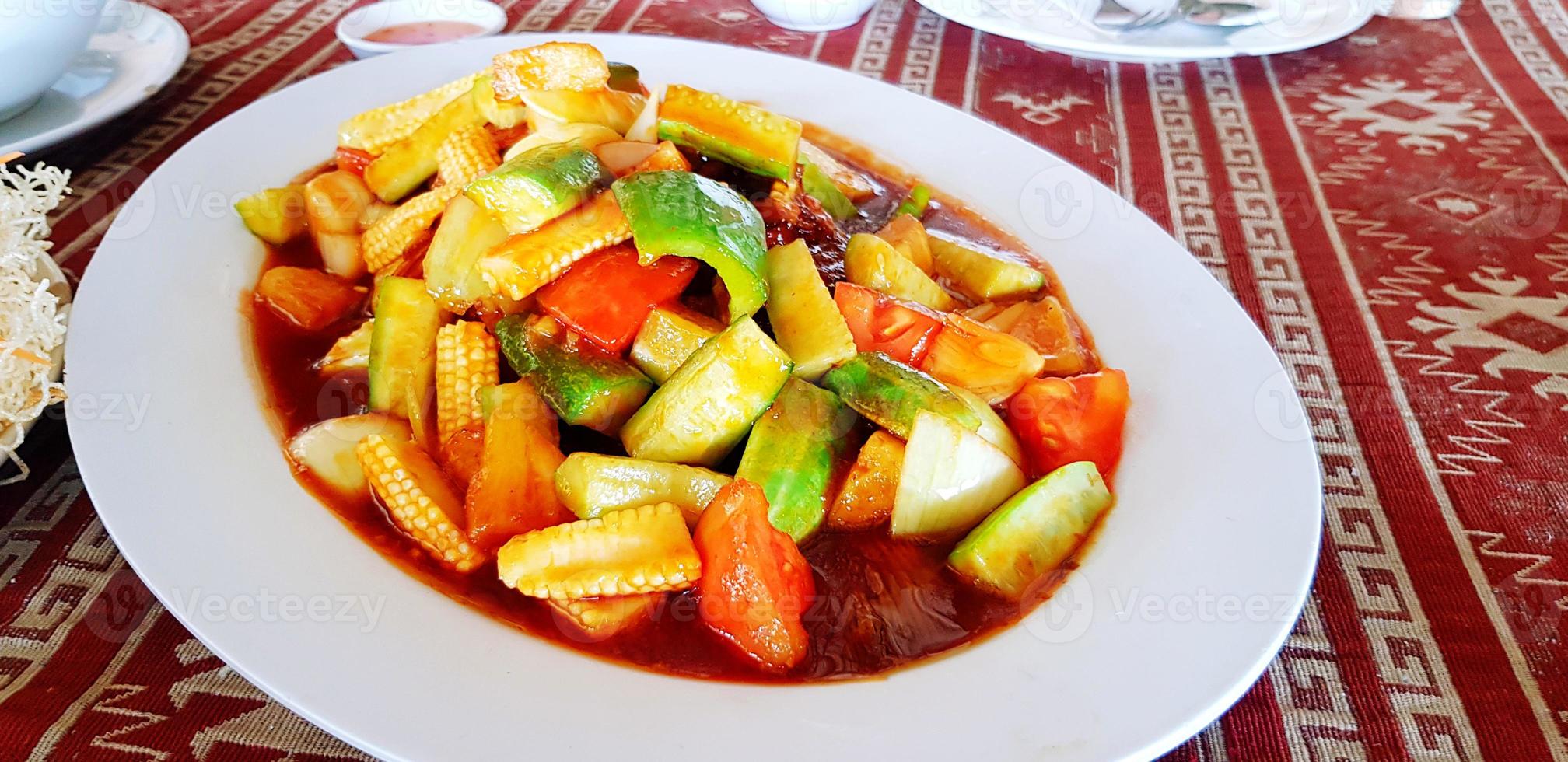 cerca arriba remover frito con rebanado pepino, maíz, tomate y agrio o dulce salsa en blanco lámina. asiático comida con Fresco vegetales en plato en rojo estera con Copiar espacio. sano estilo de vida comiendo foto