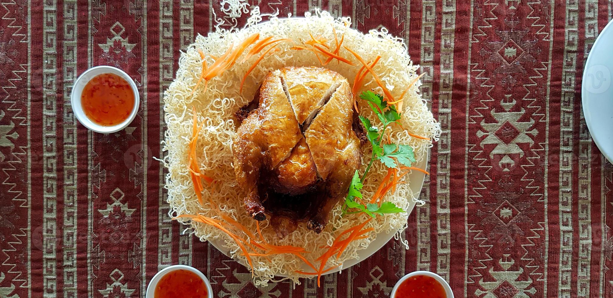Top view of baked whole chicken with fried rice noodles, sliced carrot, coriander and sweet or sour sauce in small bowl on red mat background. Flat lay of Asian food on table. Delicious meal at lunch. photo