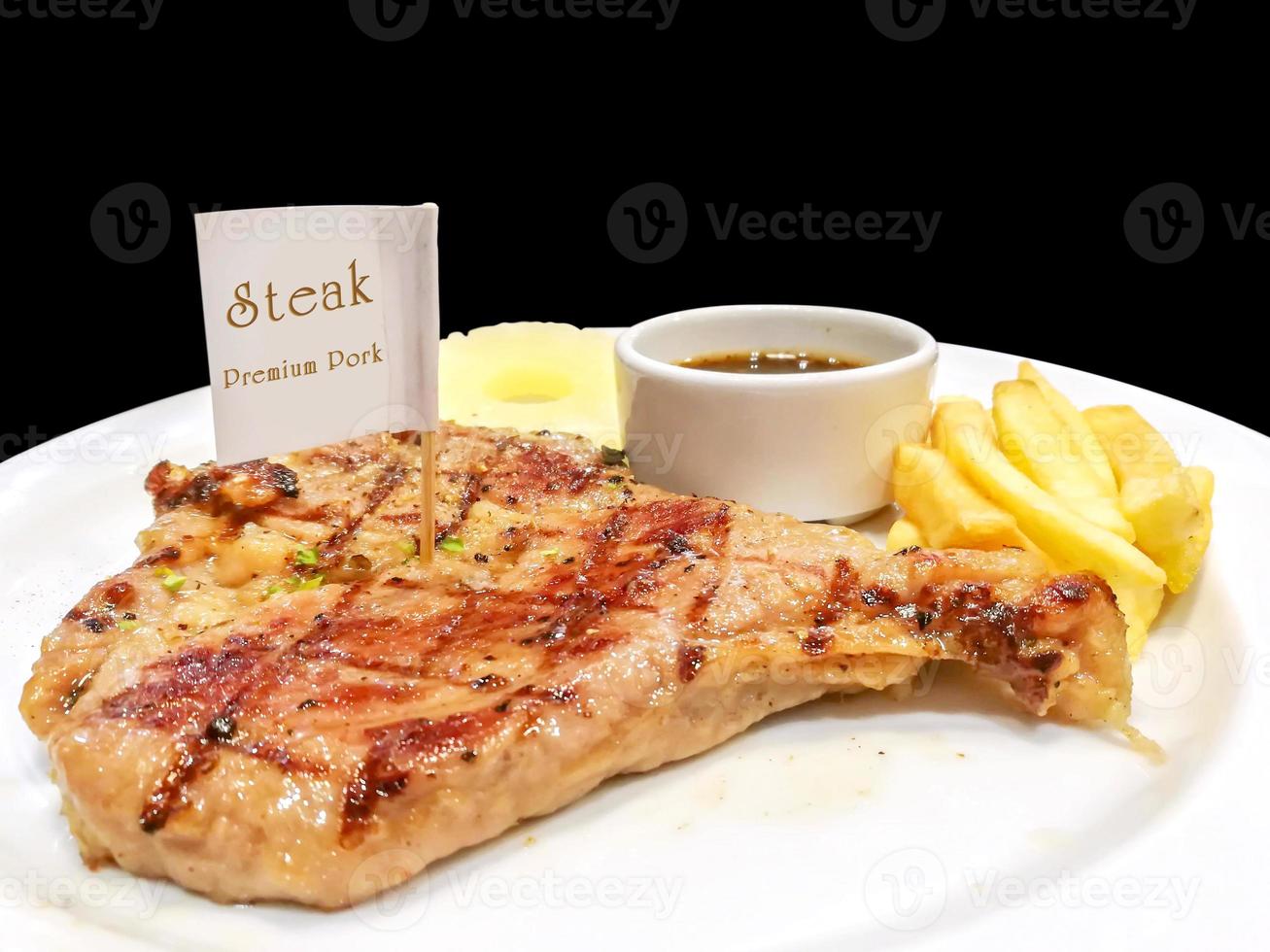 T-bone pork steak with french fries, pineapple slice and pepper sauce with steak knife and fork on white ceramic plate isolate on black background and make with paths. photo