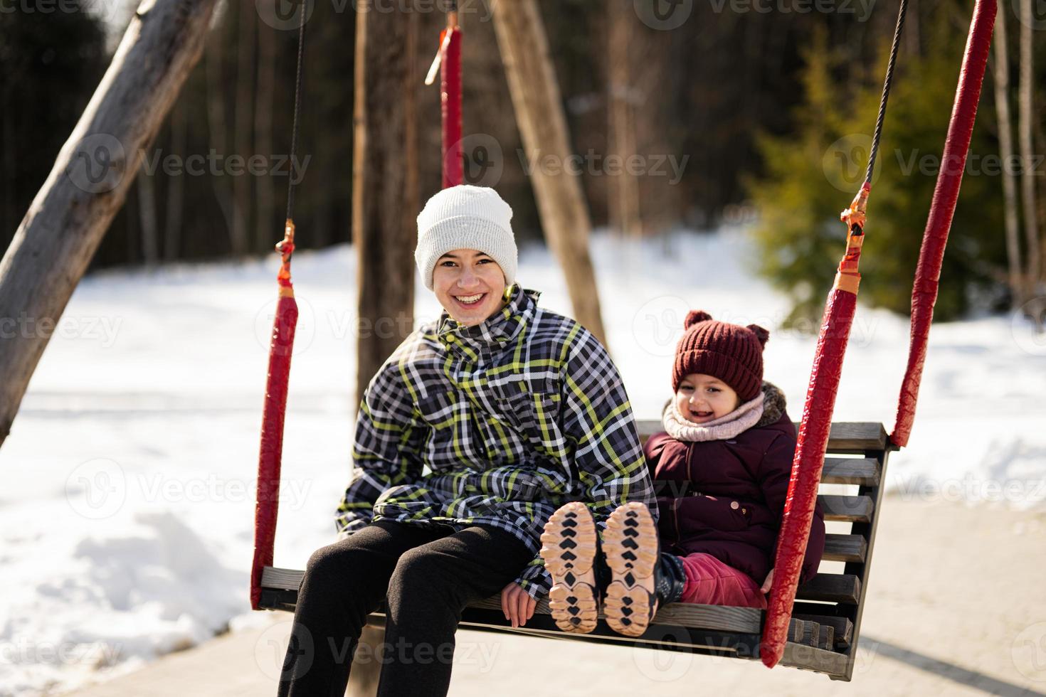 Older brother with younger sister swinging on big wooden swing in forest of mountains. photo