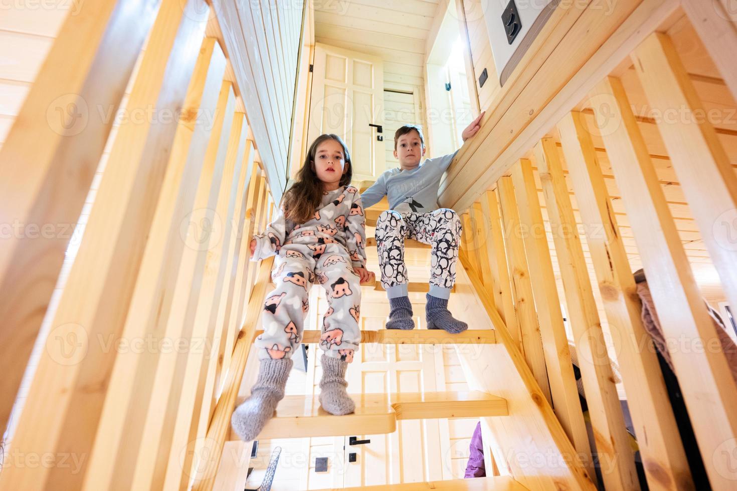 Brother with sister in pajama sit on stairs in cozy wooden tiny cabin house. Life in countryside. photo