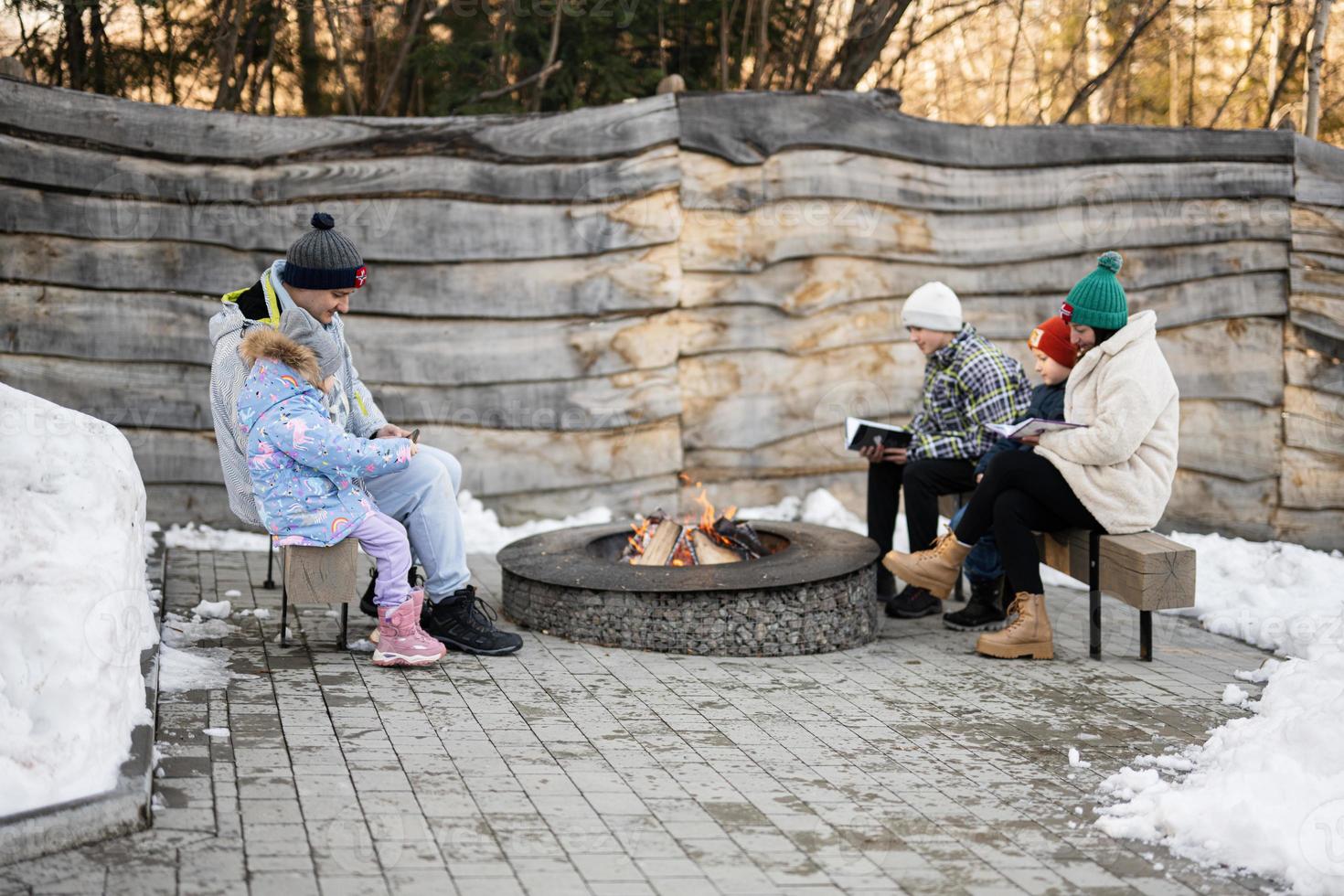 Family with three kids sitting by camp bonfire and read books on winter in forest. Children in countryside. photo