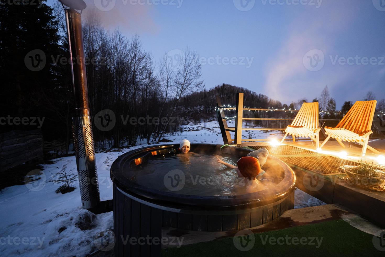 Kids enjoying bathing in wooden barrel hot tub in the terrace of the cottage. Scandinavian bathtub with a fireplace to burn wood and heat water. photo