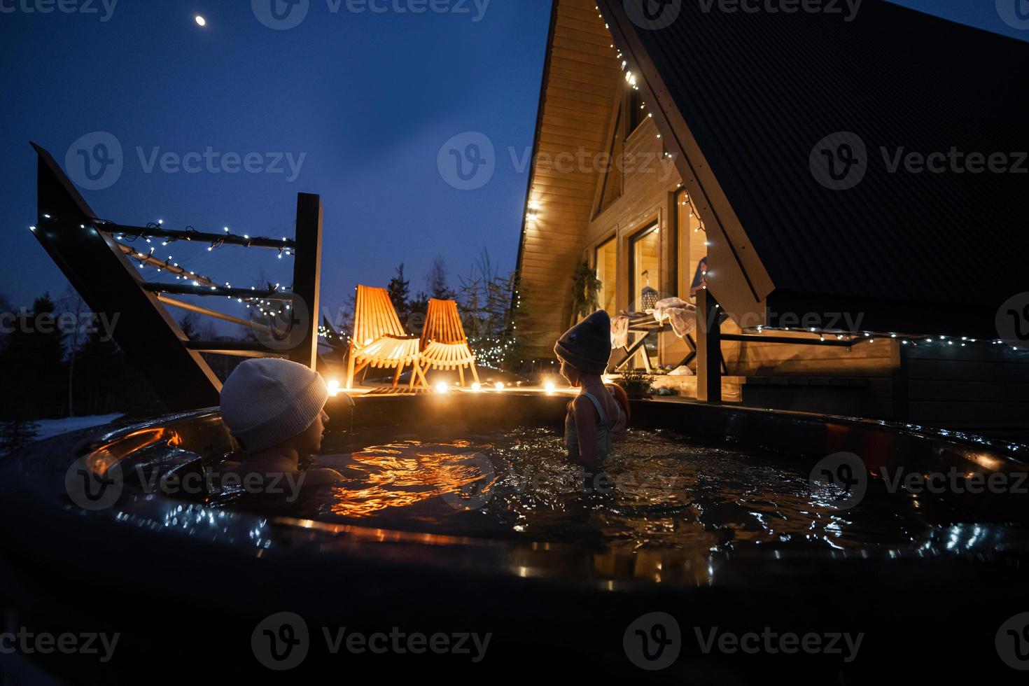 Kids enjoying bathing in wooden barrel hot tub in the terrace of the cottage. Scandinavian bathtub with a fireplace to burn wood and heat water. photo