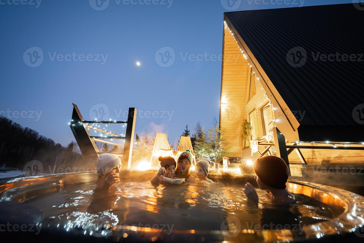 Family enjoying bathing in wooden barrel hot tub in the terrace of the cottage. Scandinavian bathtub with a fireplace to burn wood and heat water. photo