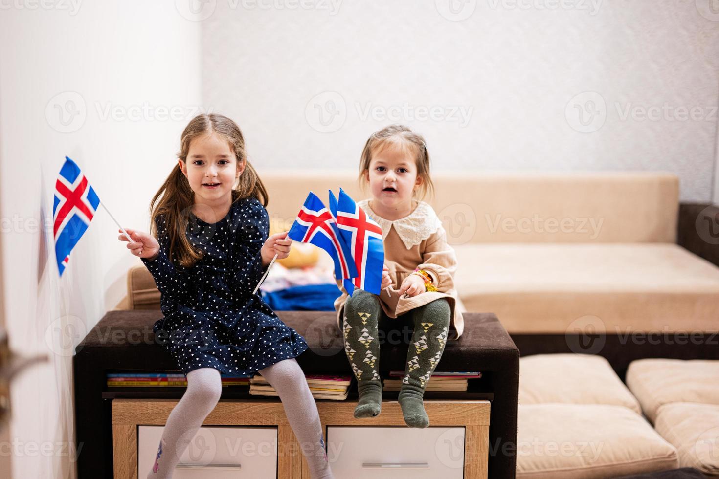 Two sisters are sitting on a couch at home with icelandic flags on hands. Iceland children girls with flag . photo