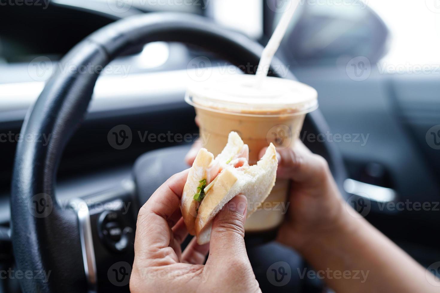 Asian woman driver hold ice coffee cup and sandwich bread for eat and drink in car, dangerous and risk an accident. photo