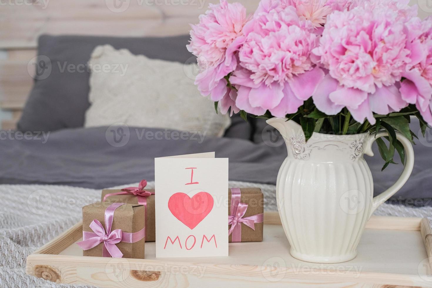 A vase of peonies and a card with the text I love mom on a tray on the bed. photo