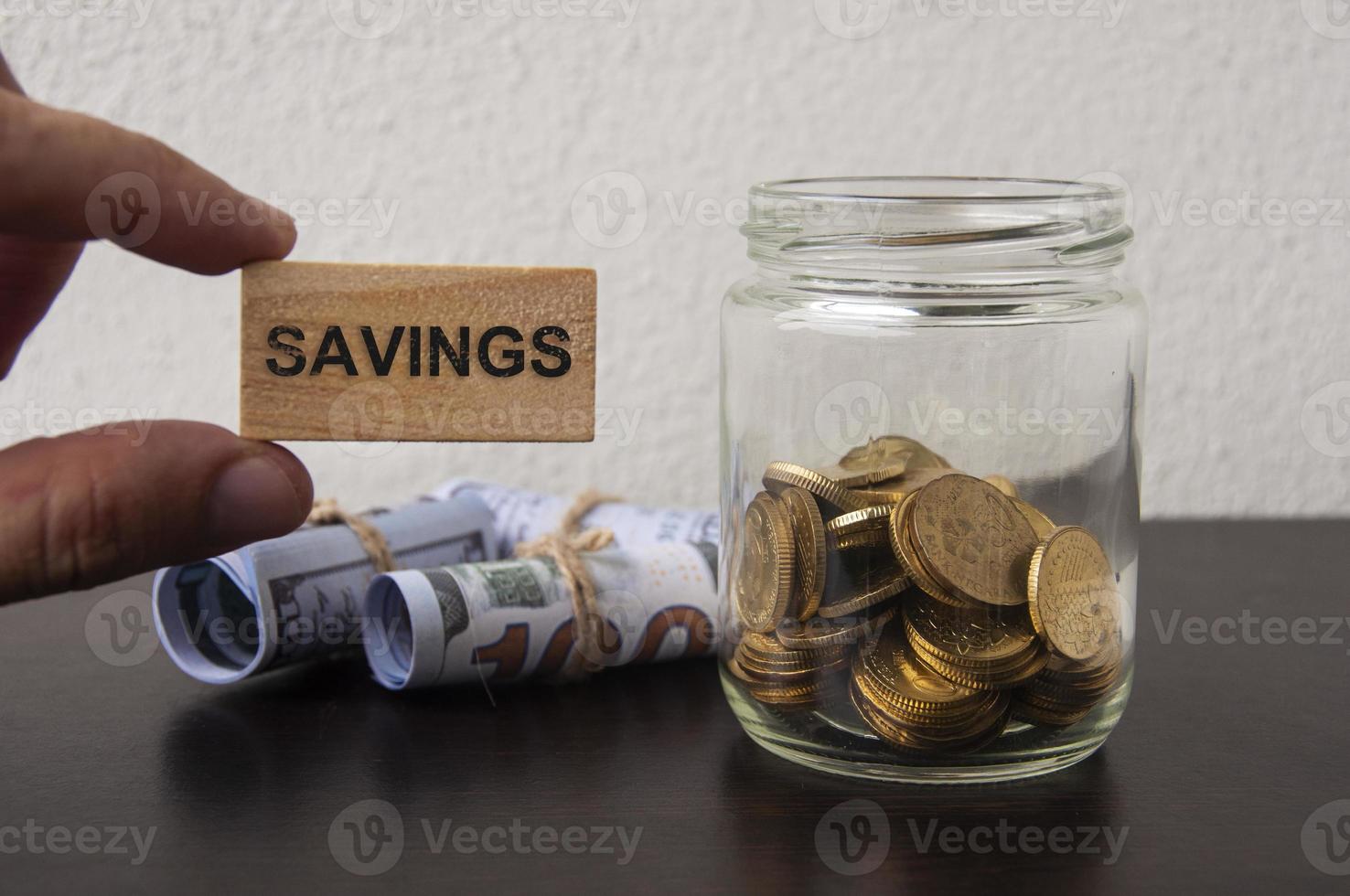 Hand holding wooden block with text Savings. Bank notes and gold coin in a jar background. photo