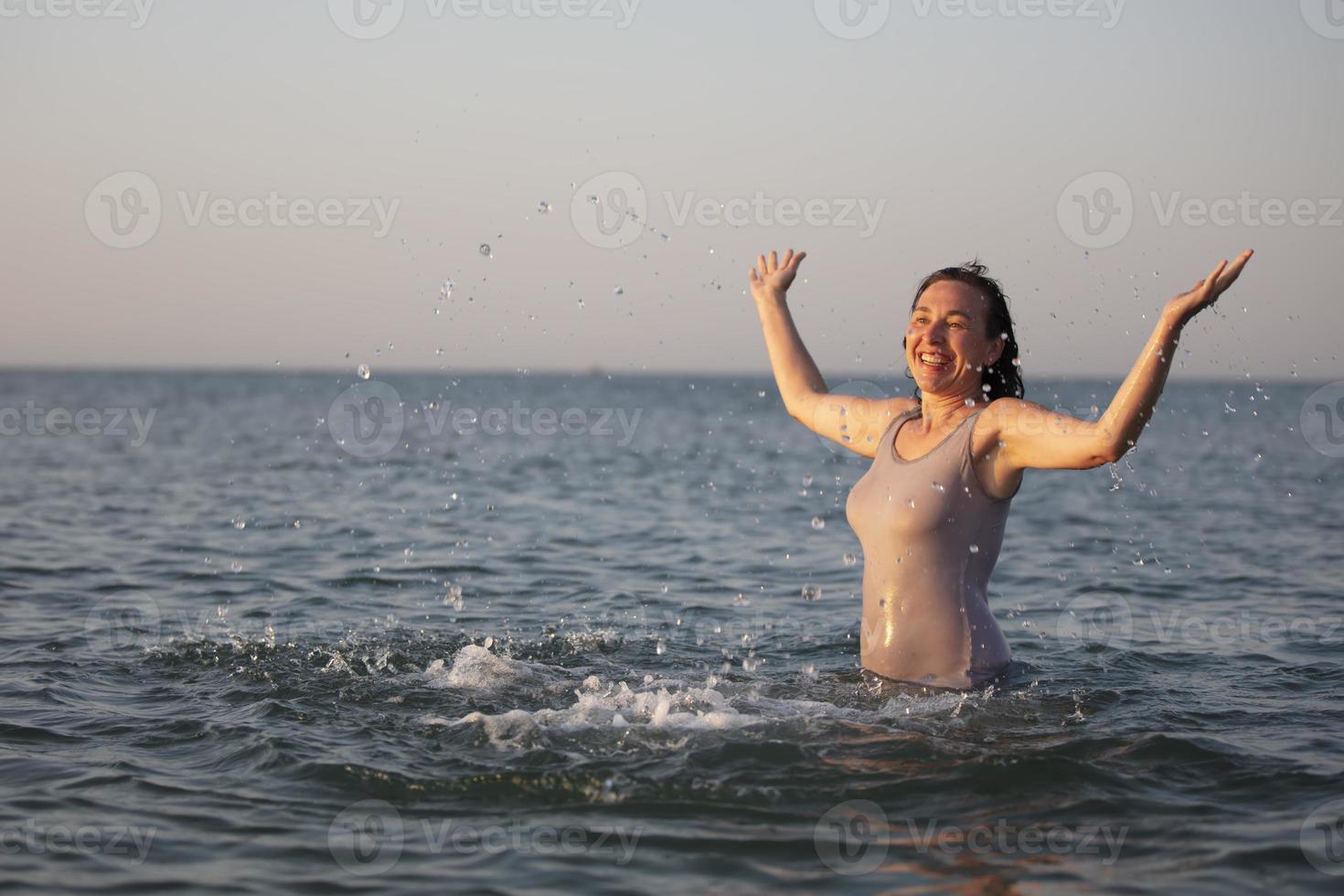 Happy woman at the sea in the sun. Woman in a swimsuit splashes water on a background of waves. Summer vacation on the beach. photo