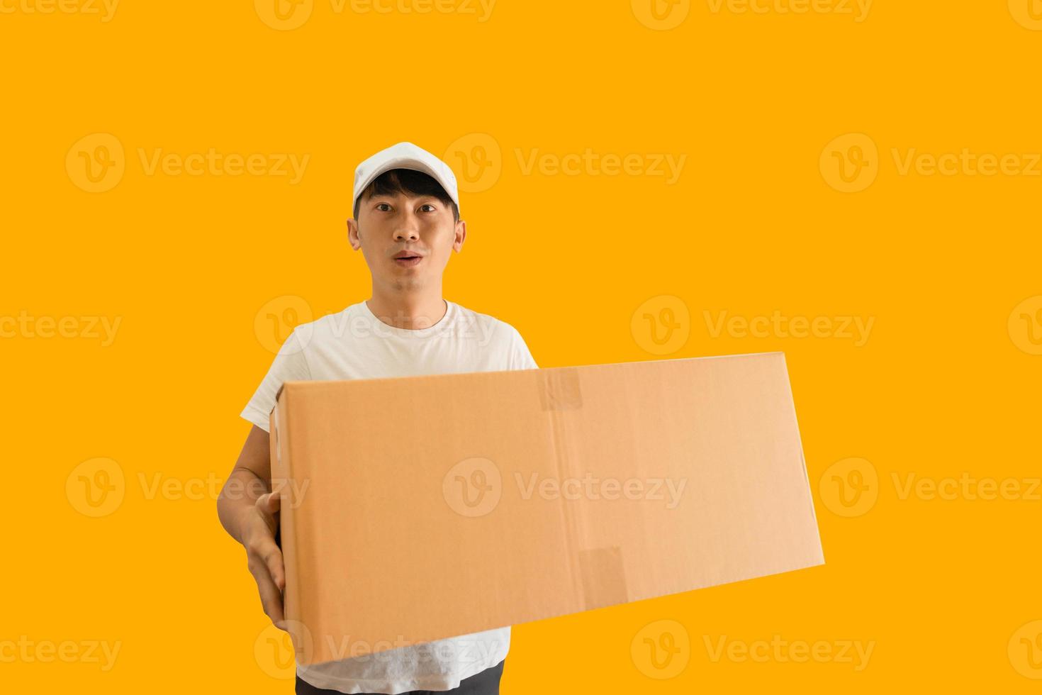 Young Asian delivery man wearing cap and white blank t-shirt holding parcel post box isolated on yellow background. express delivery service concept. photo