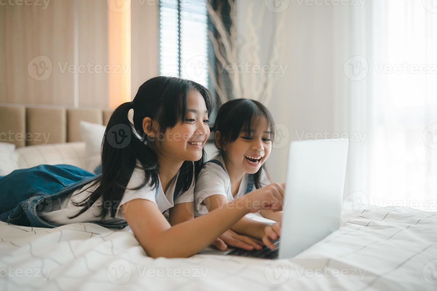 Sibling Asian girl child on the bed and learning online on a laptop Internet. Virtual class lesson on video during self isolation at home. Distant remote video education. Modern school study for kids photo