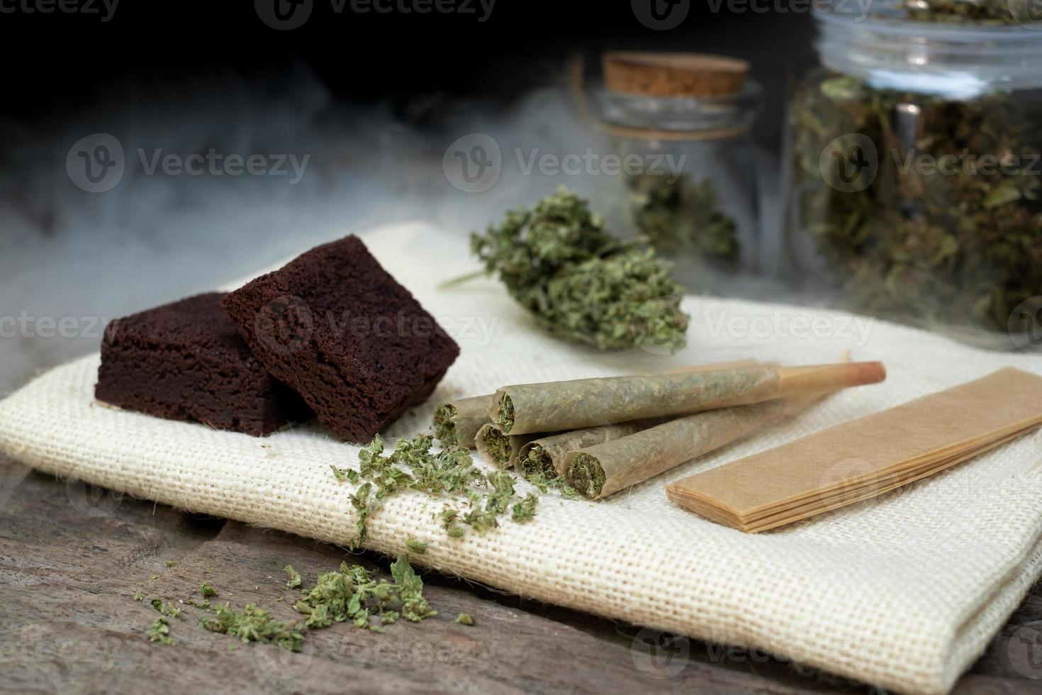 Pre-Roll cannabis joints and cake brownies with curing cannabis buds in a clear glass jar laying a black background photo