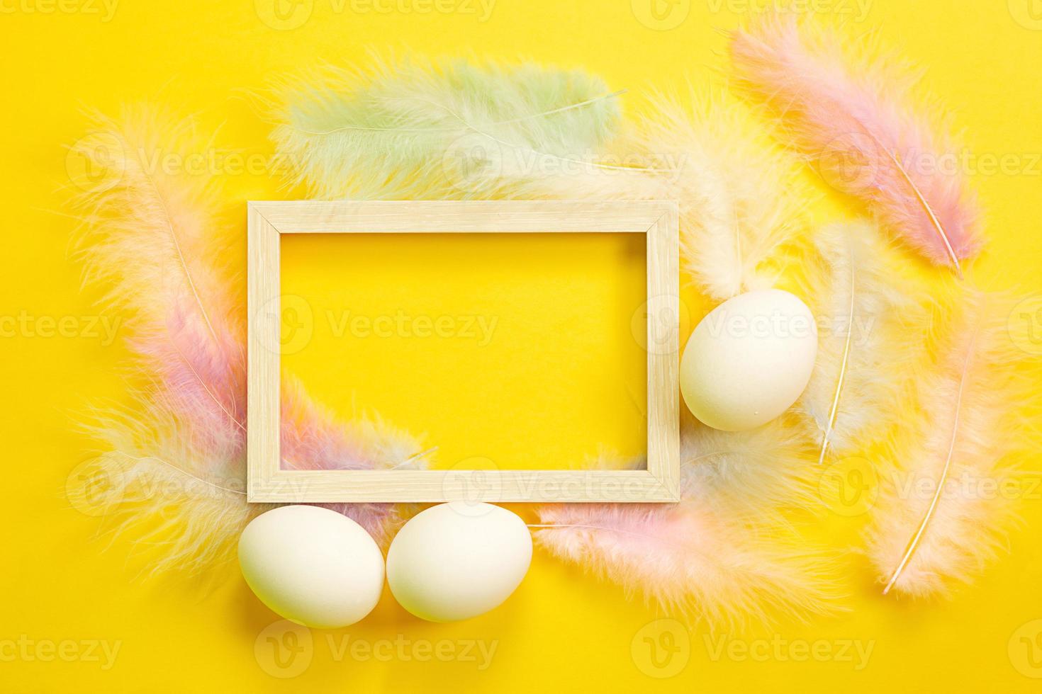 Easter white frame on a bright yellow background of chicken eggs and colored delicate feathers. Spring, religious holiday, Easter decoration, greeting, copy space, mock up photo