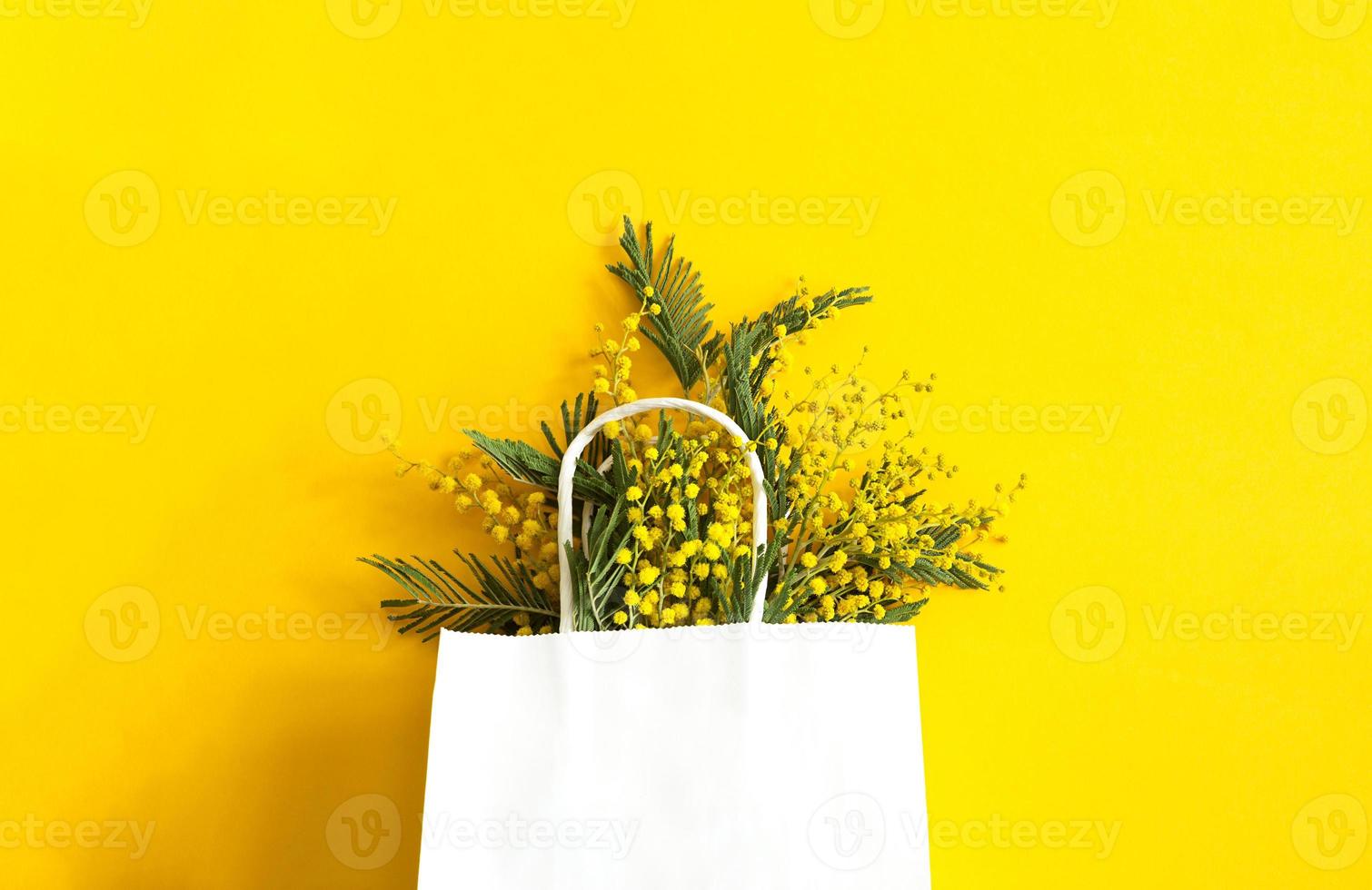 un ramo de flores de mimosa en un blanco burlarse de arriba regalo bolsa. primavera compras, regalos y promociones para internacional De las mujeres día. amarillo fondo, espacio de copia. foto
