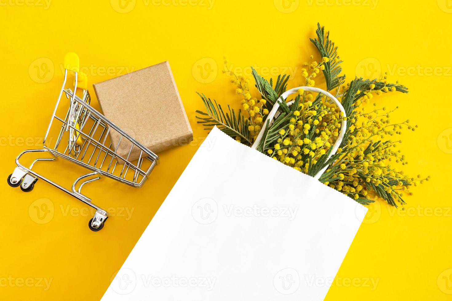 A bouquet of mimosa in a white mock up paper bag, craft gift box and a grocery cart. Spring shopping, gifts and promotions for International Women's Day. Yellow background, copyspace, flatlay. photo