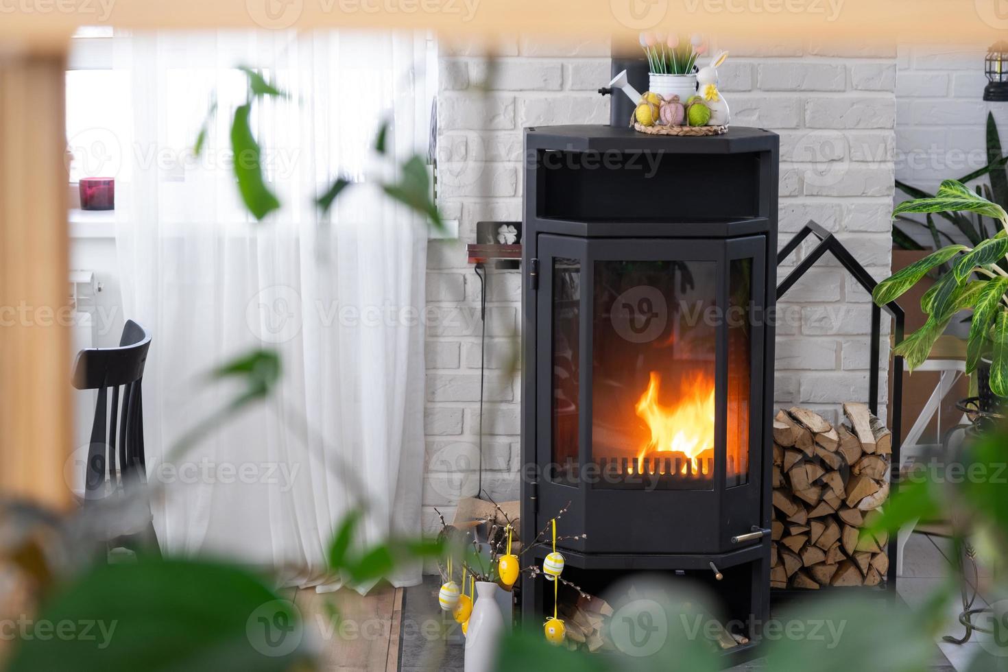 Easter decor near fireplace stove with fire and firewood. Cozy home hearth in interior with potted plans, colorful easter eggs, easter bunny and bouquet. Spring in a country house photo