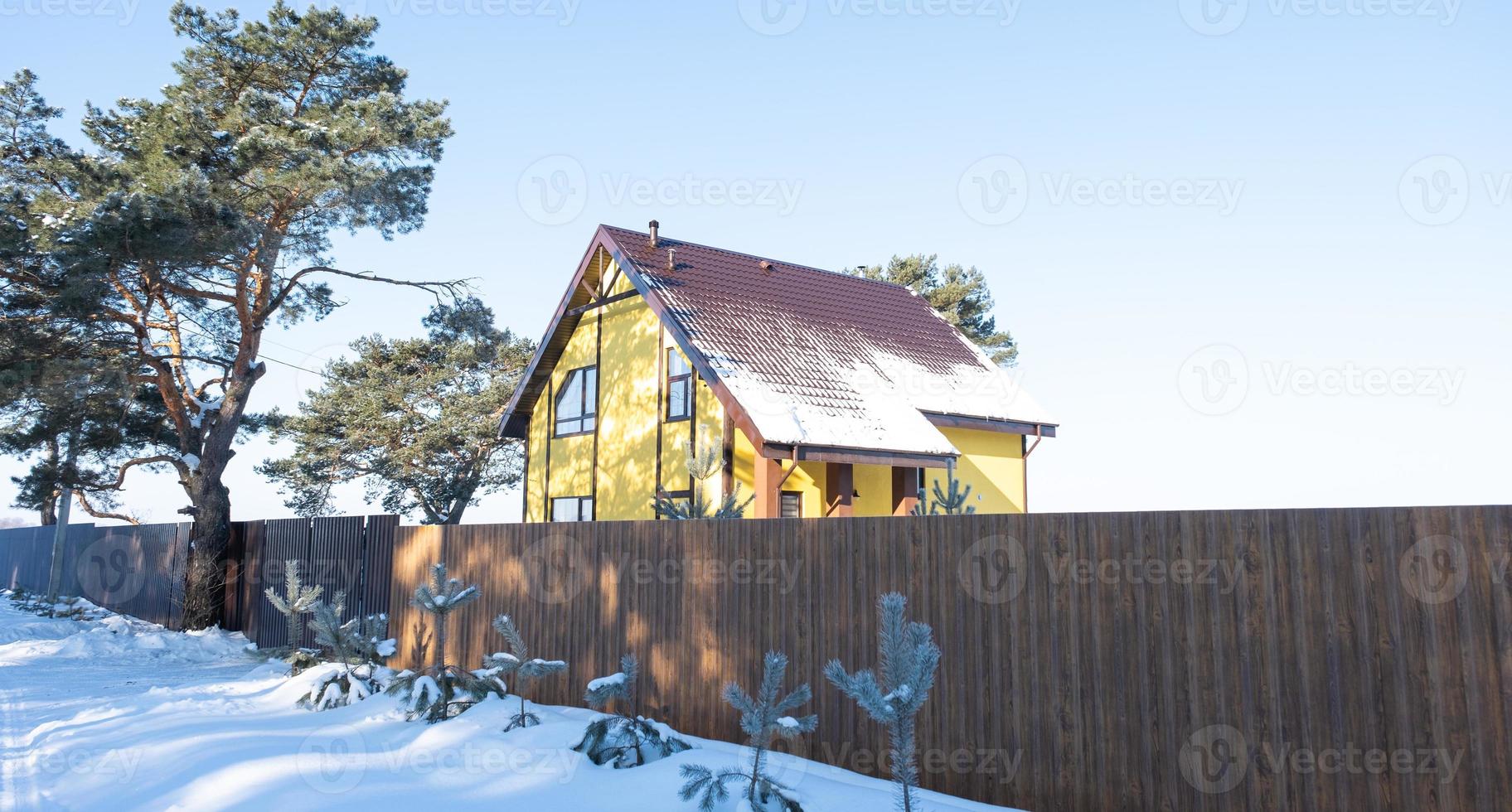 A yellow cozy house in the snow in winter in the village is surrounded by pine trees. Snow-covered roof, heating and ventilation pipes, trapezoid windows photo