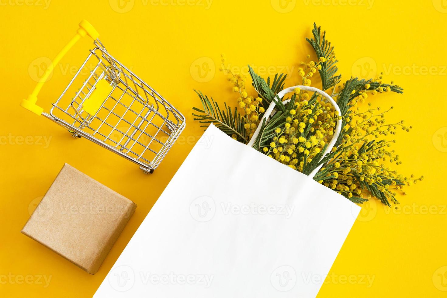 A bouquet of mimosa in a white mock up paper bag, craft gift box and a grocery cart. Spring shopping, gifts and promotions for International Women's Day. Yellow background, copyspace, flatlay. photo