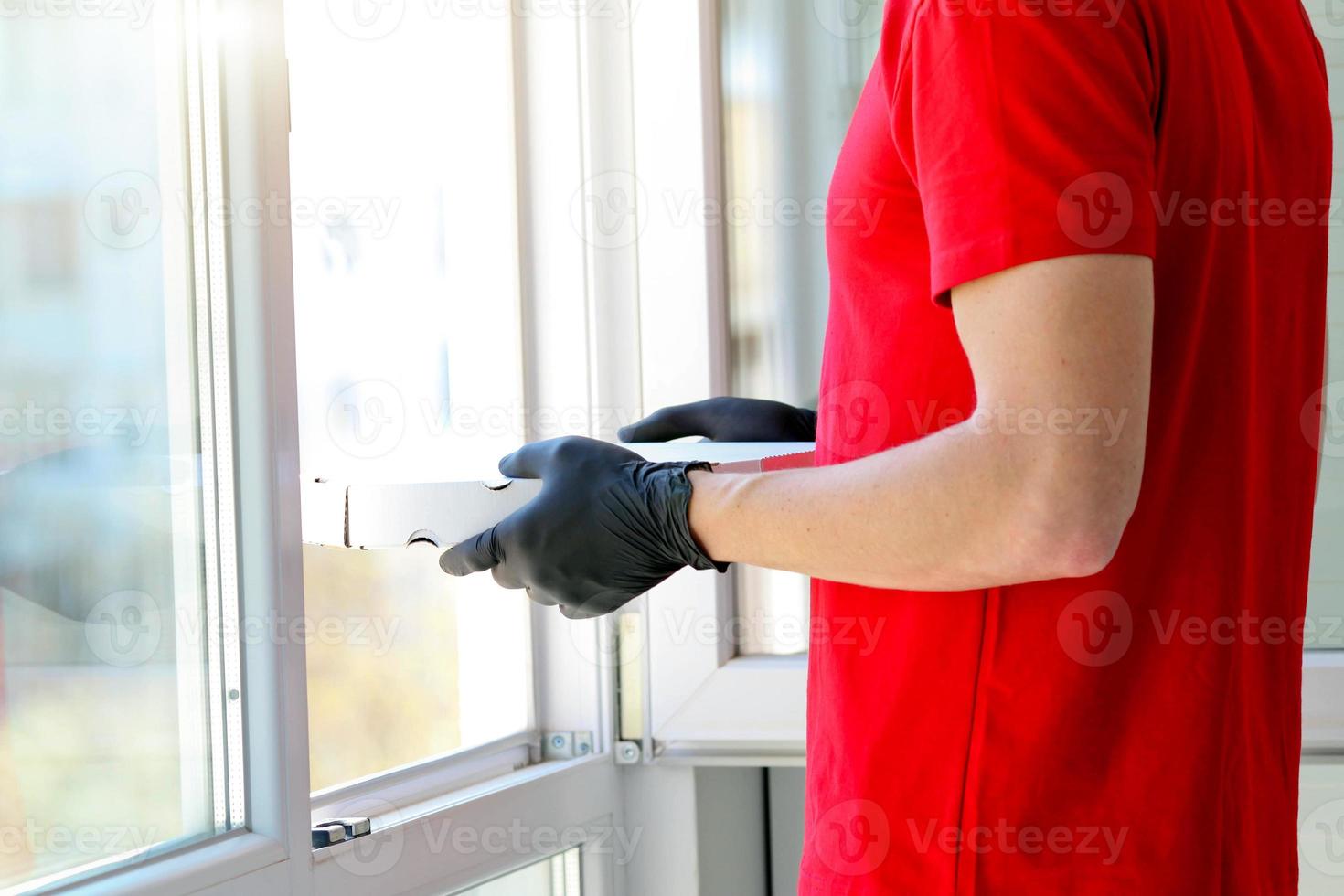 un hombre manos caucho guantes con un caja Pizza cerca el ventana . mensajero. comida entrega durante el cuarentena de el coronavirus pandemia. emisión de pedidos, en línea orden. foto