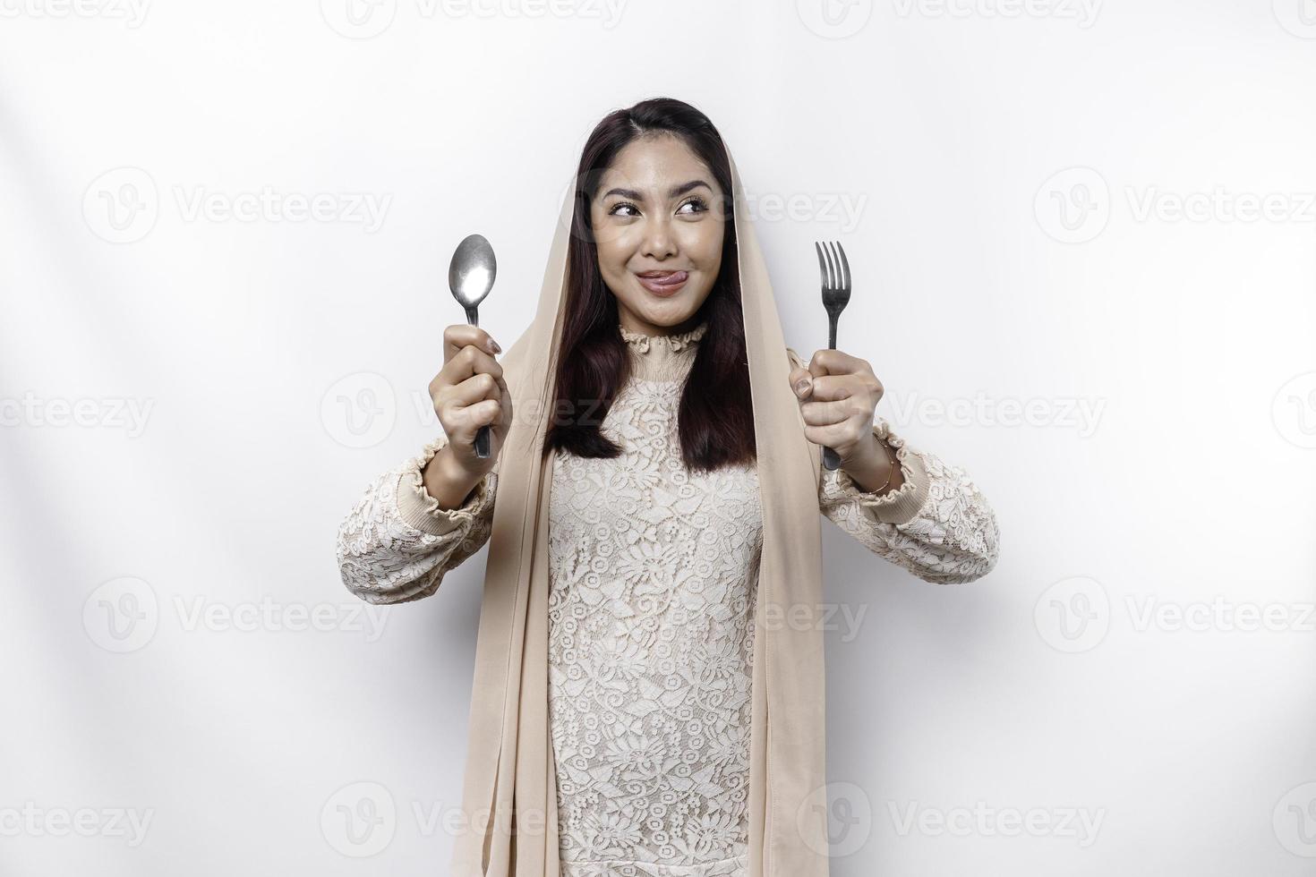 An Asian Muslim woman is fasting and hungry and holding utensils cutlery while looking aside thinking about what to eat photo