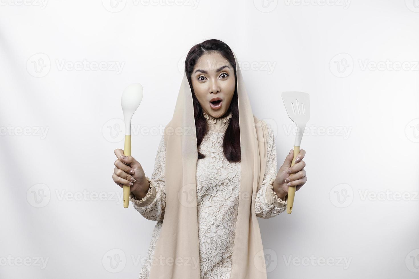 Excited Asian Muslim woman wearing a headscarf, holding cooking ware and smiling, isolated by white background photo