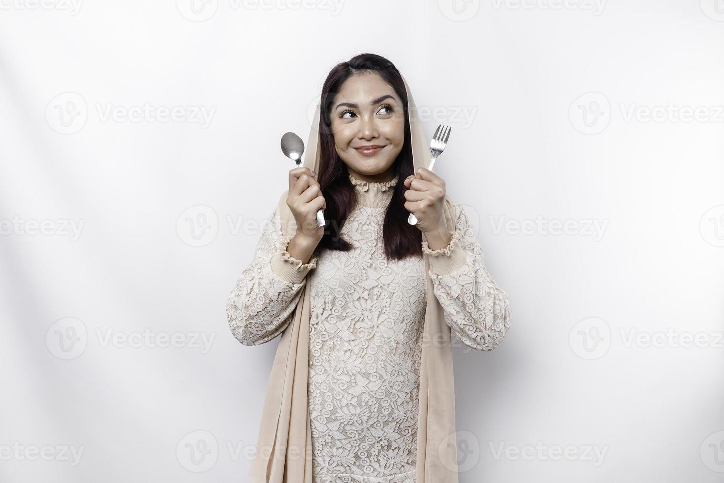 An Asian Muslim woman is fasting and hungry and holding utensils cutlery while looking aside thinking about what to eat photo