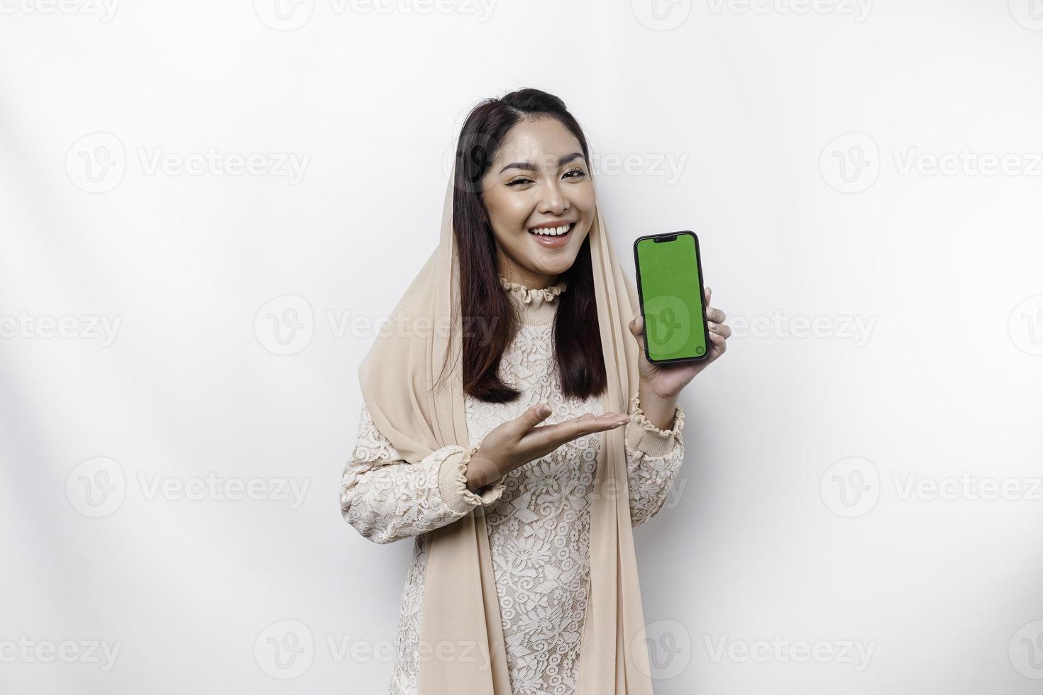 A portrait of a happy Asian Muslim woman wearing a headscarf, showing her phone screen, isolated by white background photo