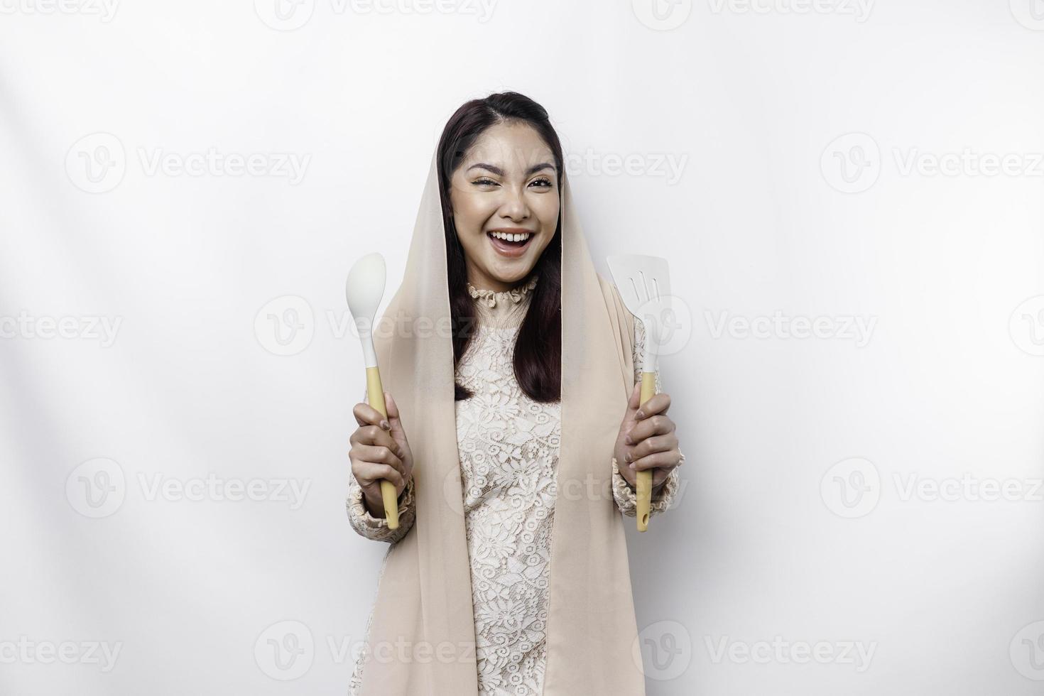 Excited Asian Muslim woman wearing a headscarf, holding cooking ware and smiling, isolated by white background photo