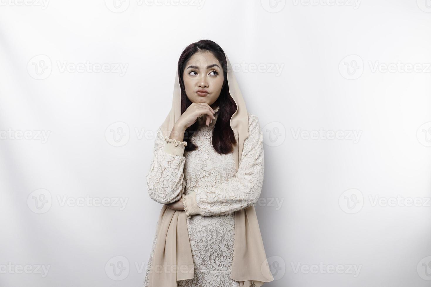 A thoughtful young Asian Muslim woman wearing headscarf while looking aside, isolated by a white background photo