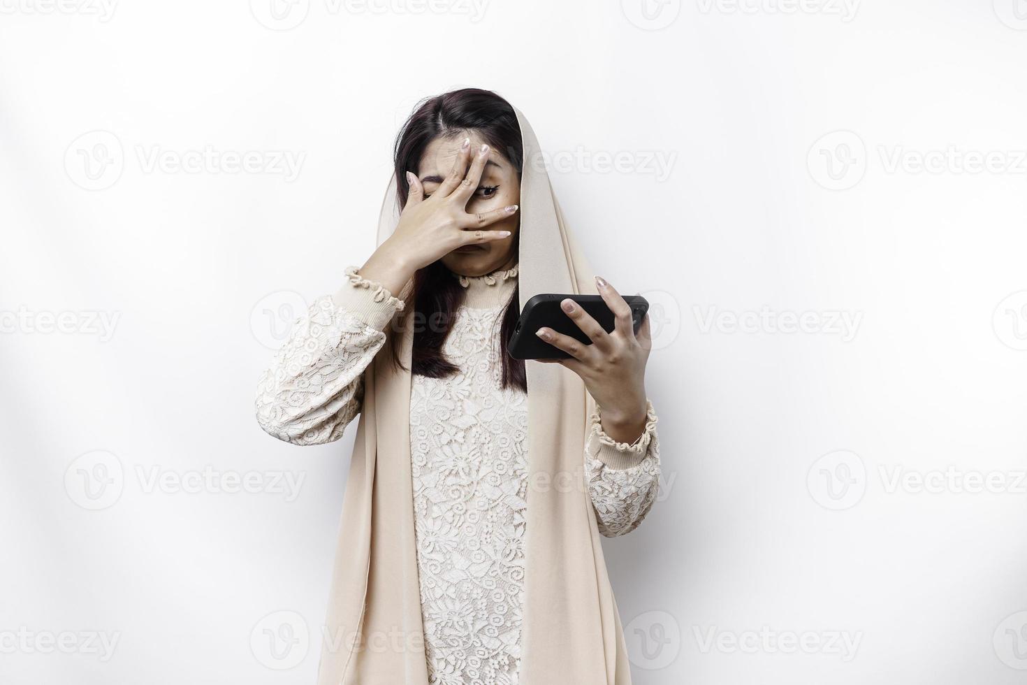 A funny Asian Muslim woman wearing a headscarf is peeking at inappropriate content on her phone isolated on white background. photo