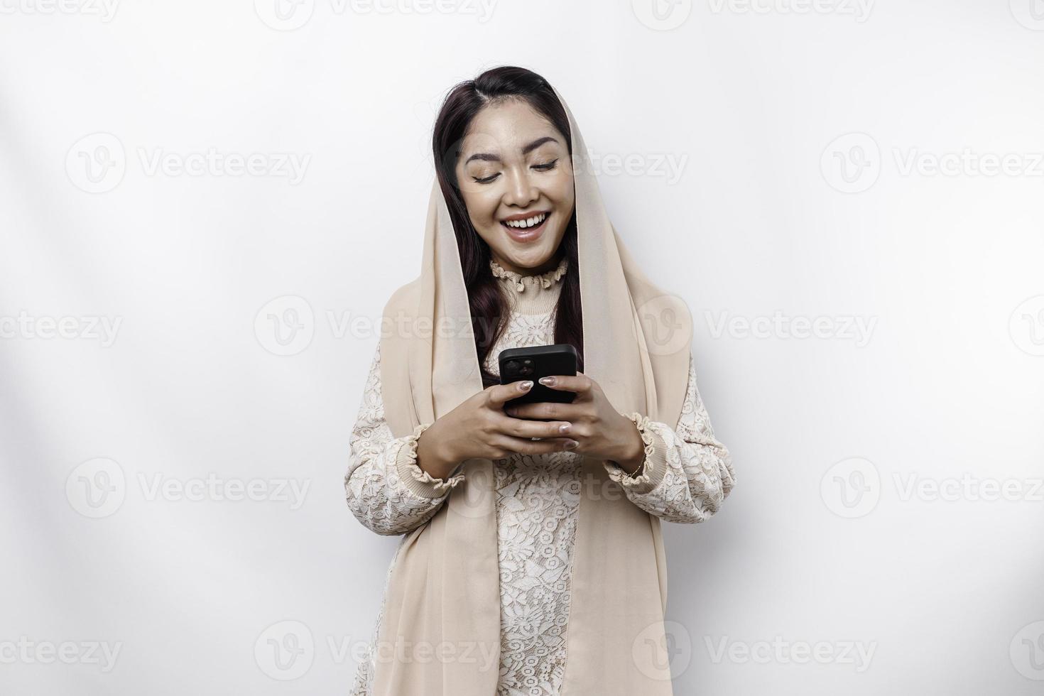 A portrait of a happy Asian Muslim woman wearing a headscarf, holding her phone, isolated by white background photo