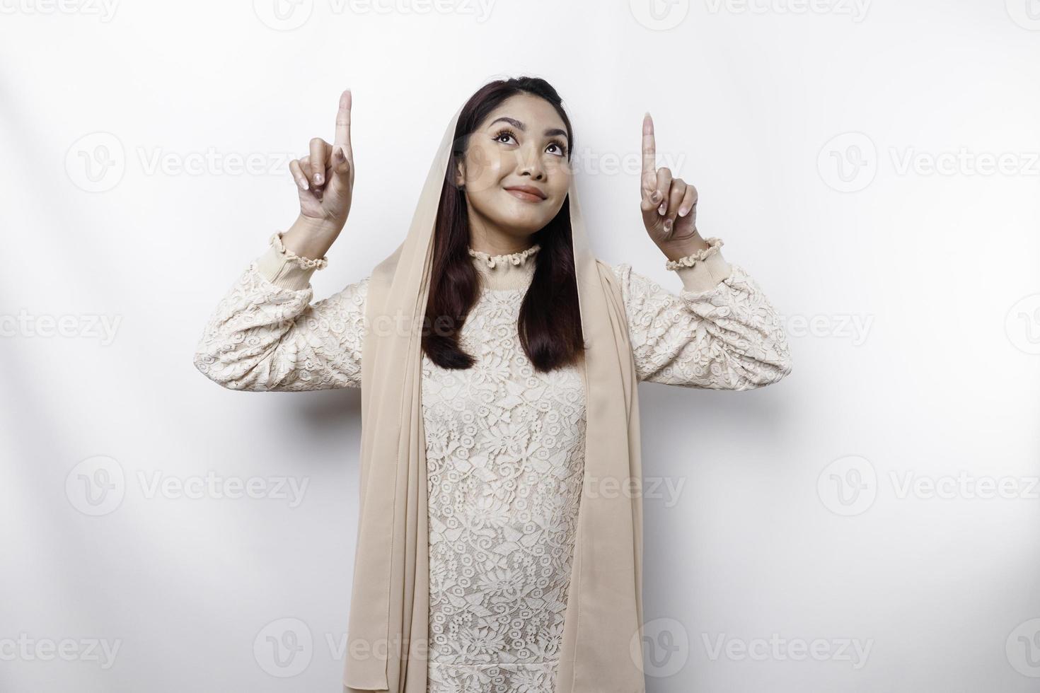 Excited Asian Muslim woman wearing hijab pointing at the copy space above her, isolated by white background photo