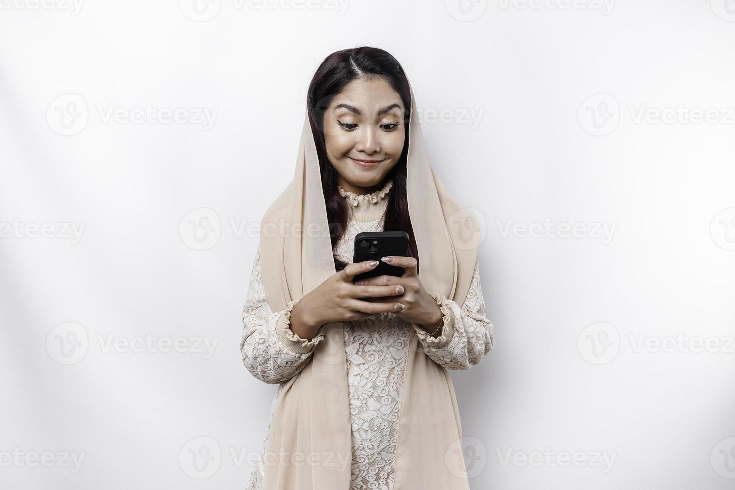 A portrait of a happy Asian Muslim woman wearing a headscarf, holding her phone, isolated by white background photo